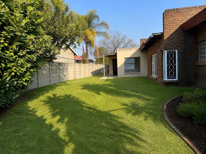 A rear view of the property featuring a brick facade, a small covered patio area, and a well-maintained lawn enclosed by a boundary wall.