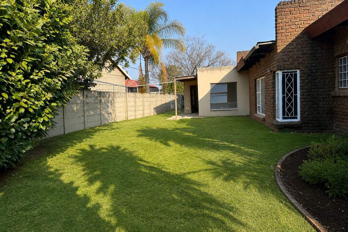 A rear view of the property featuring a brick facade, a small covered patio area, and a well-maintained lawn enclosed by a boundary wall.