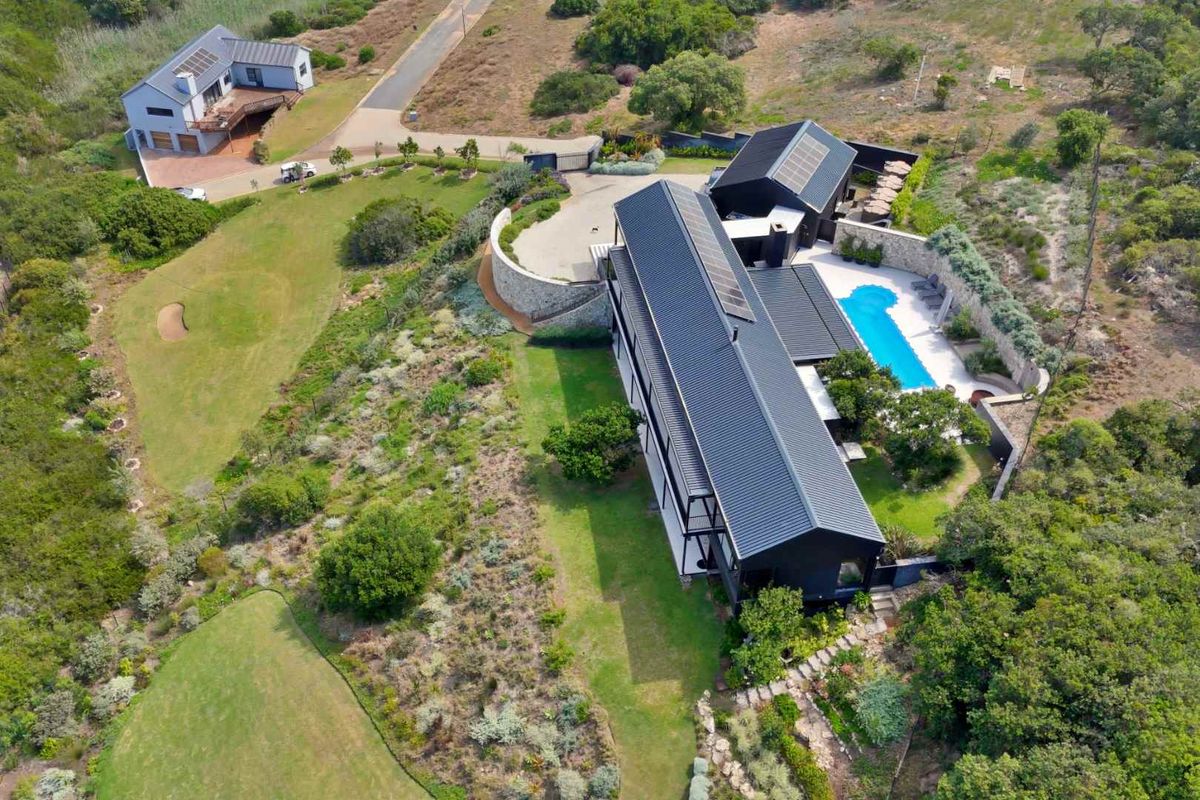 Aerial view of a modern black-clad residence featuring a swimming pool, landscaped gardens, and solar panels, situated in a scenic, open estate setting.