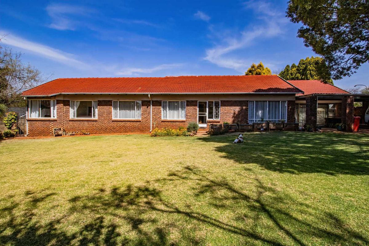 A rear view of a single-story brick house featuring a tiled roof, large garden lawn, and a covered patio area.