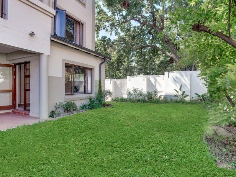 A private rear garden featuring a well-maintained lawn, mature trees, and a tiled patio area adjacent to the apartment building with secure perimeter fencing.
