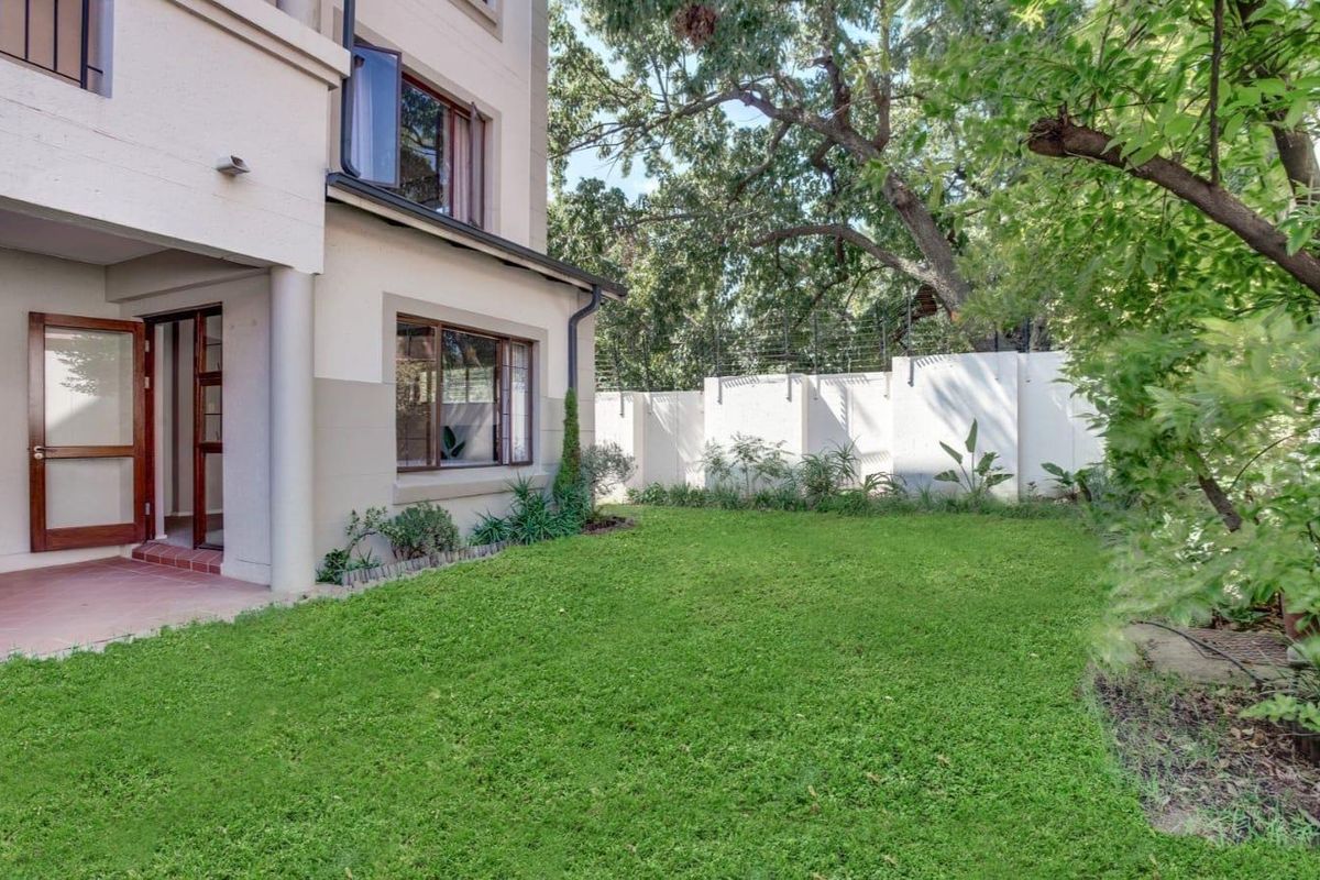 A private rear garden featuring a well-maintained lawn, mature trees, and a tiled patio area adjacent to the apartment building with secure perimeter fencing.