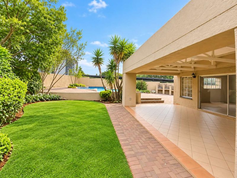 Spacious covered patio featuring tiled flooring, overlooking a well-maintained garden, swimming pool, and built-in outdoor braai area.