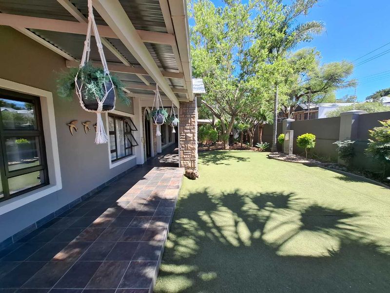 A covered patio area featuring tiled flooring and a stone-clad pillar, overlooking a well-maintained garden with artificial lawn and boundary wall.