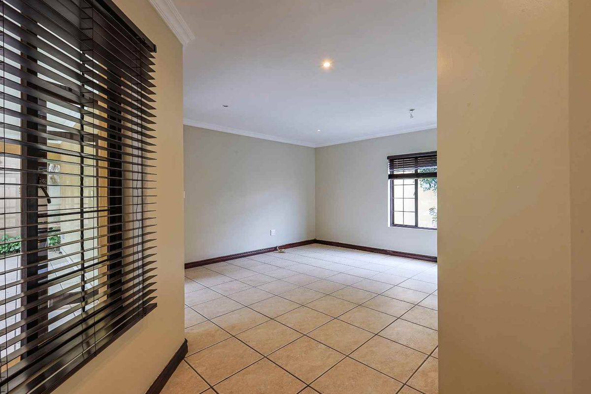 A bright, empty living room featuring neutral tiled flooring, recessed downlighting, and dark-framed windows with fitted blinds.