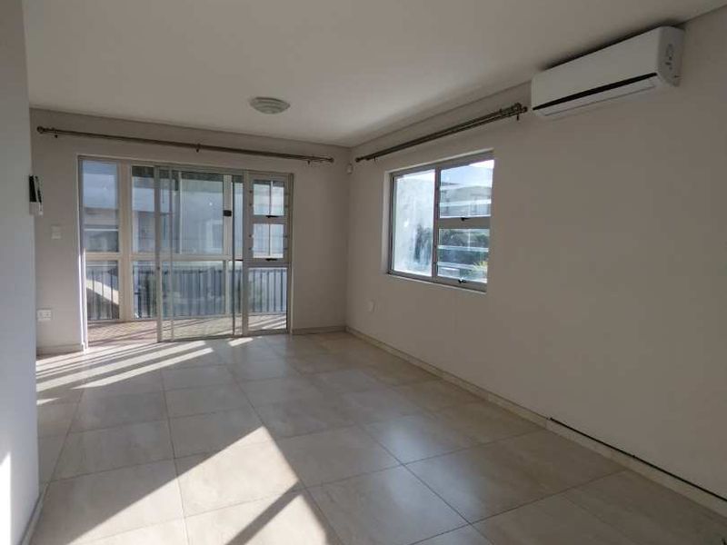 A bright, empty living area featuring large format floor tiles, a wall-mounted air conditioning unit, and sliding glass doors leading to an outdoor space.