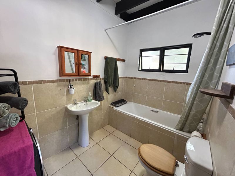 A functional bathroom featuring a pedestal basin, built-in bathtub with shower, and tiled finishes, complemented by exposed ceiling beams and a louver window.