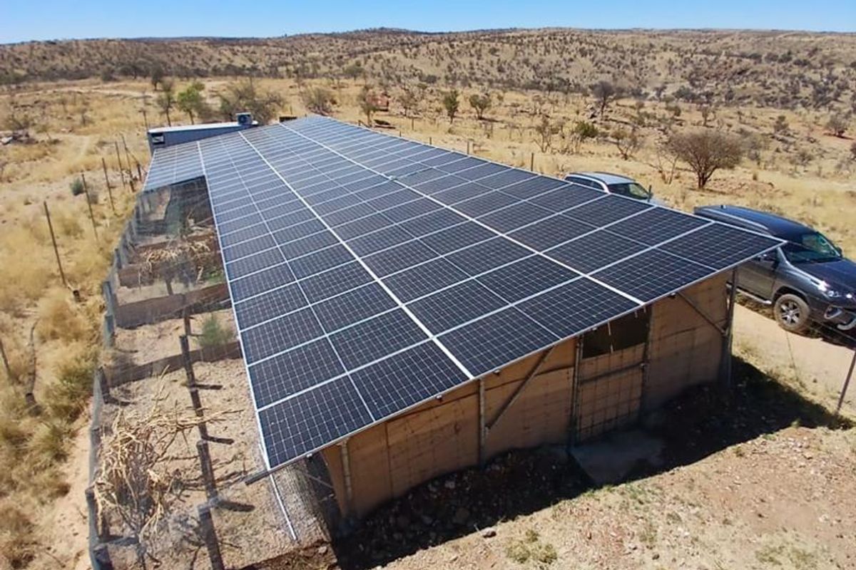 Aerial view of a large solar panel installation mounted on a metal structure with an adjacent fenced enclosure on a rural small holding.