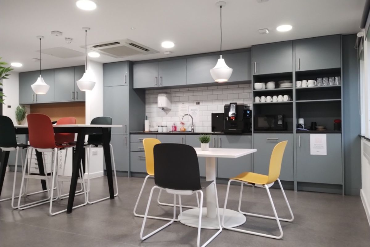 A modern office kitchen area featuring sleek grey cabinetry, subway tile backsplash, recessed lighting, and integrated sink facilities.