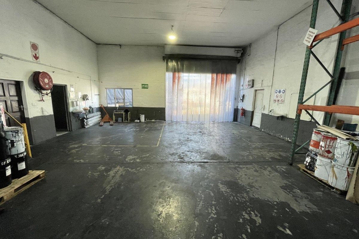 Spacious industrial warehouse unit featuring high ceilings, concrete flooring, a large roller shutter door with PVC strip curtains, and integrated storage racking.