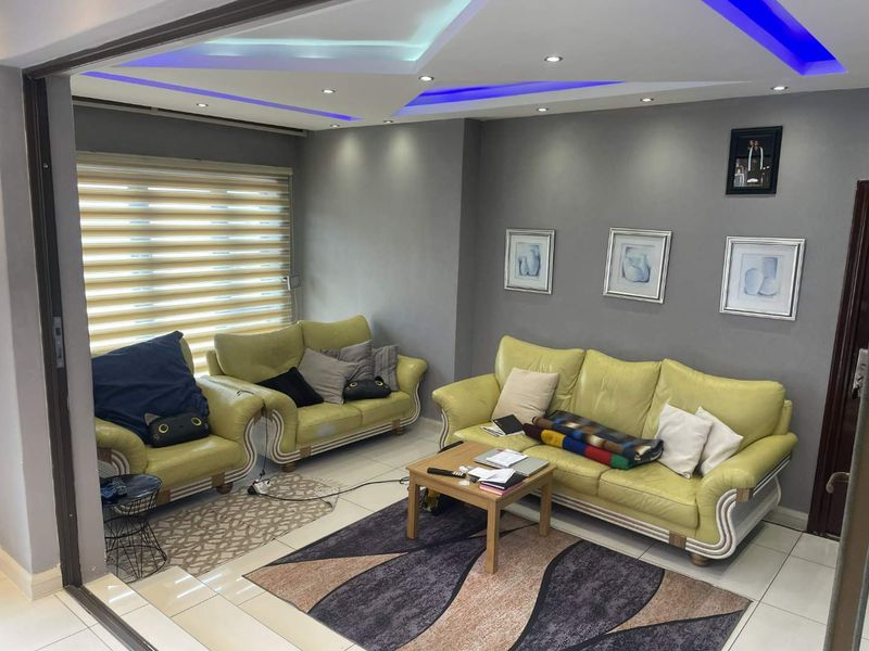 A modern living room featuring tiled flooring, recessed ceiling lights with blue LED accents, and large windows with fitted roller blinds.