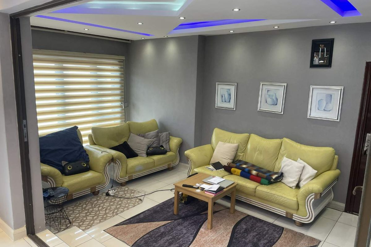 A modern living room featuring tiled flooring, recessed ceiling lights with blue LED accents, and large windows with fitted roller blinds.