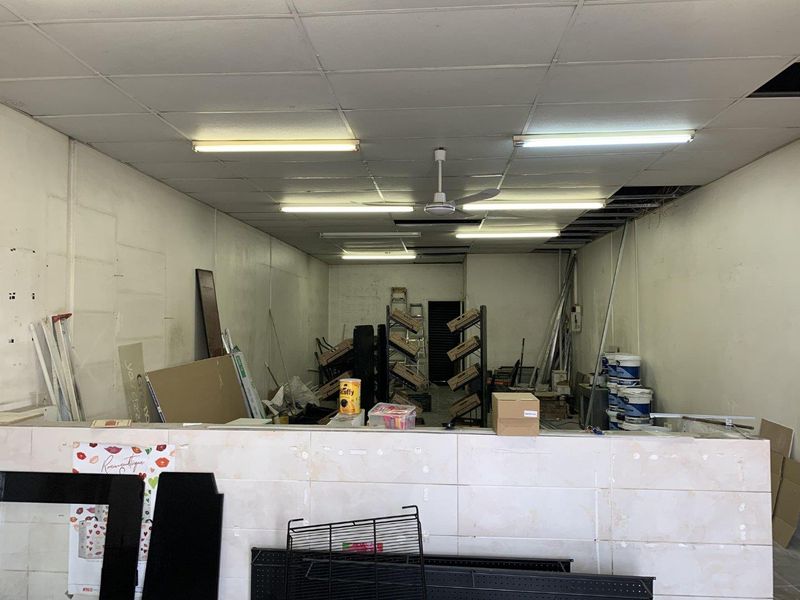 An open-plan retail space featuring a tiled service counter, suspended ceiling with fluorescent lighting, and a ceiling fan, currently in need of refurbishment.