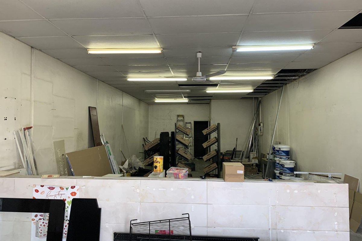 An open-plan retail space featuring a tiled service counter, suspended ceiling with fluorescent lighting, and a ceiling fan, currently in need of refurbishment.