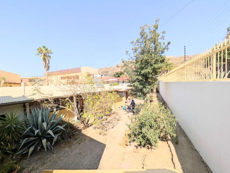 Rear exterior view of the property featuring a concrete pathway, perimeter wall with security fencing, and an unlandscaped garden area.