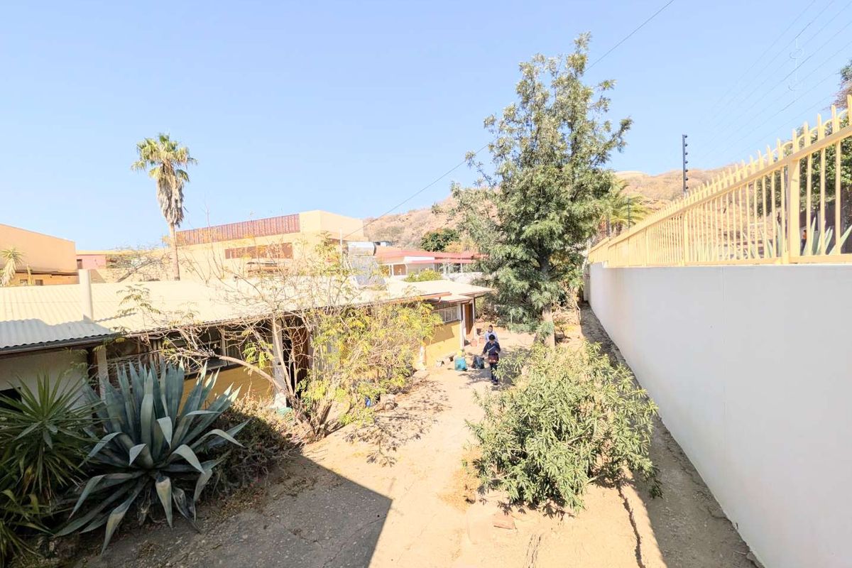 Rear exterior view of the property featuring a concrete pathway, perimeter wall with security fencing, and an unlandscaped garden area.