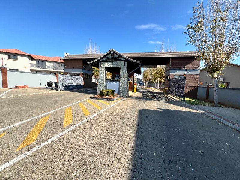 A well-maintained residential estate entrance featuring a brick security gatehouse, paved driveway, and automated boom gate access.