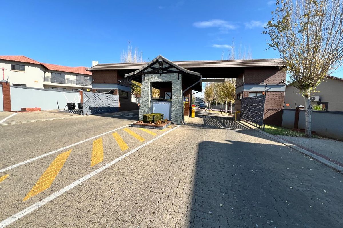 A well-maintained residential estate entrance featuring a brick security gatehouse, paved driveway, and automated boom gate access.