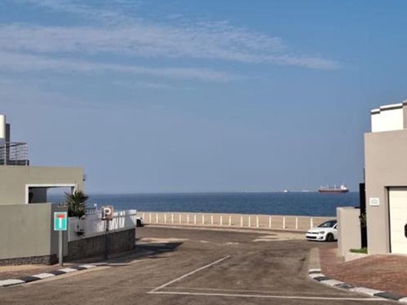 A street view of the townhouse complex approach, featuring paved roads, boundary walls, and a clear view of the ocean in the distance.