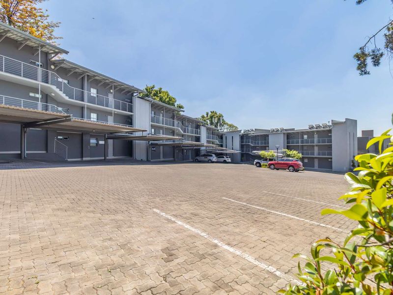 Exterior view of a multi-story apartment complex featuring a large paved parking area with covered bays and accessible balcony walkways.
