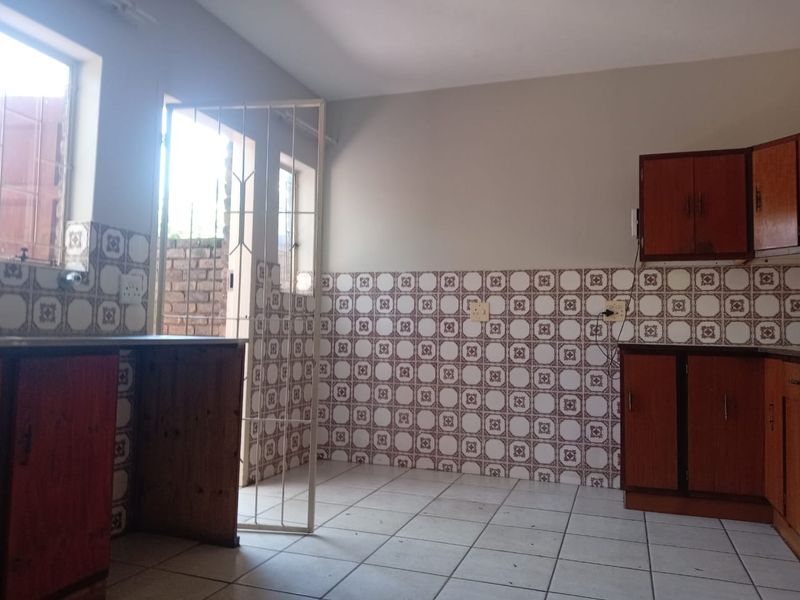 An empty kitchen featuring dated wooden cabinetry, patterned tiled splashbacks, and tiled flooring with direct access to an exterior area via a security gate.