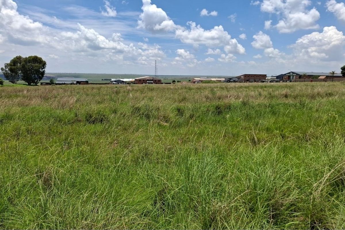 A wide, open expanse of vacant residential land featuring natural grass coverage with distant views of surrounding commercial and industrial structures.