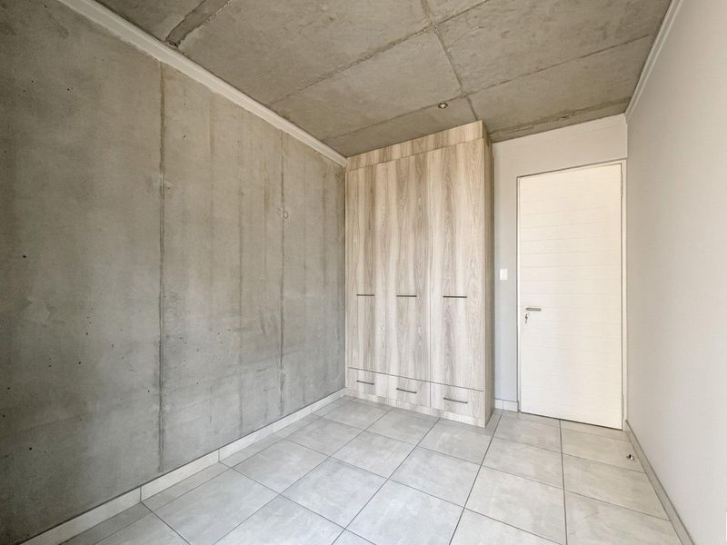 A modern bedroom featuring an exposed concrete ceiling and wall, neutral tiled flooring, and a built-in wooden wardrobe.