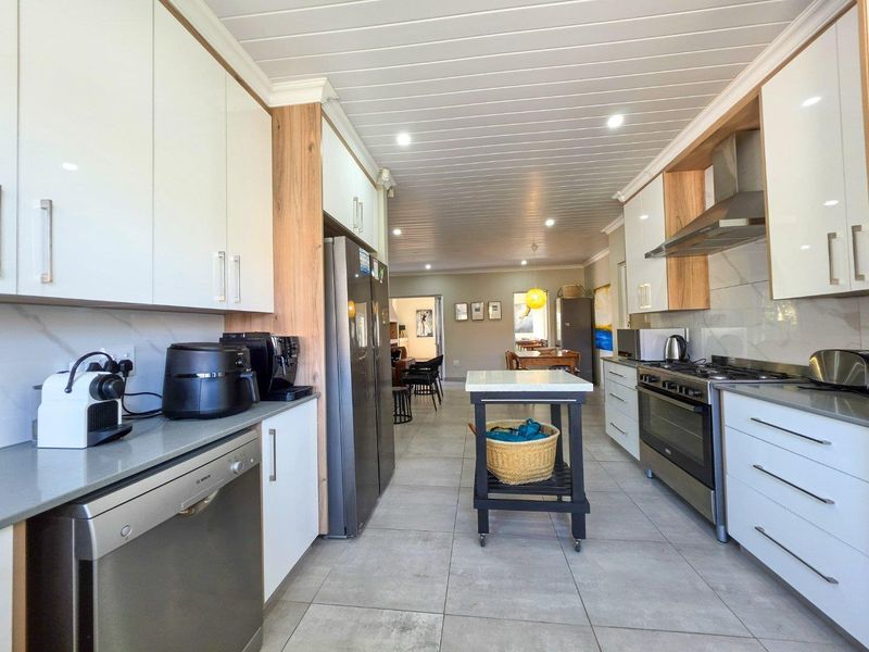 Modern galley-style kitchen featuring sleek white cabinetry, stone countertops, high-end stainless steel appliances, and tiled flooring, leading into an open-plan living area.