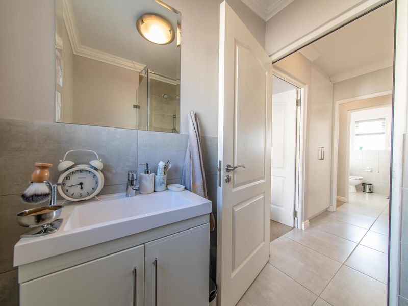 A clean, modern bathroom featuring a vanity cabinet with basin, wall-mounted mirror, and neutral tiled flooring, leading into a hallway.