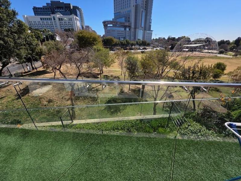 A modern balcony featuring a glass balustrade with a stainless steel handrail and artificial turf flooring, overlooking a park and urban skyline.