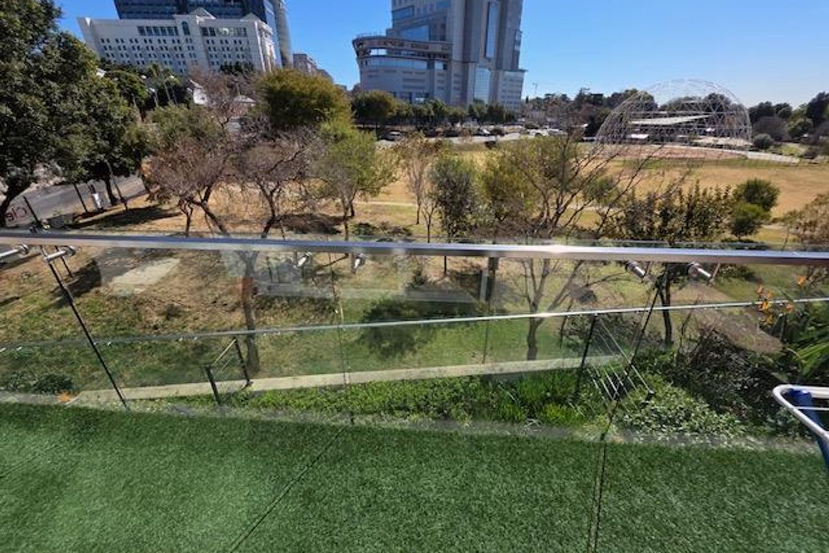 A modern balcony featuring a glass balustrade with a stainless steel handrail and artificial turf flooring, overlooking a park and urban skyline.