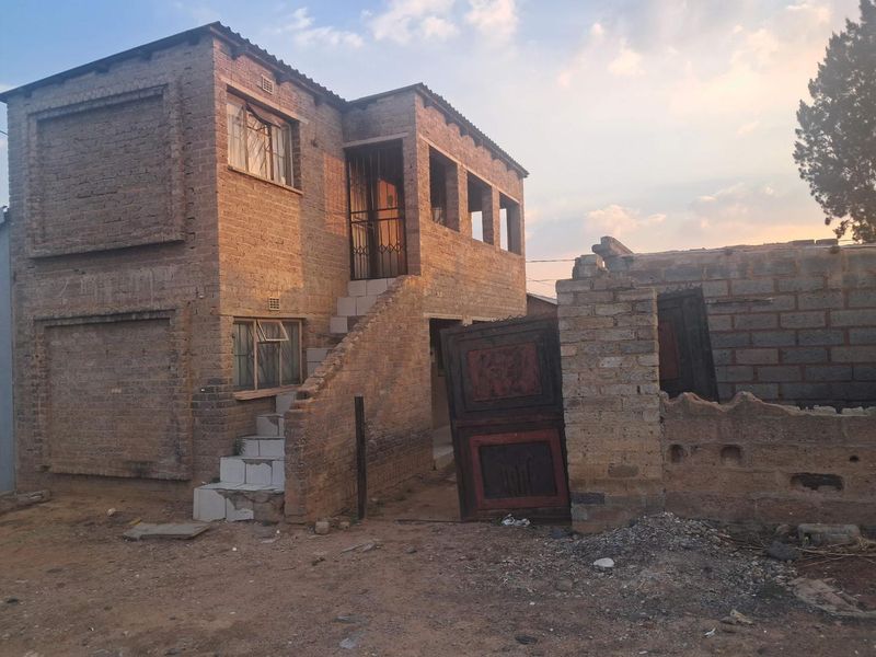 Two-story exposed brick house featuring an external staircase and a damaged boundary wall with a broken gate.