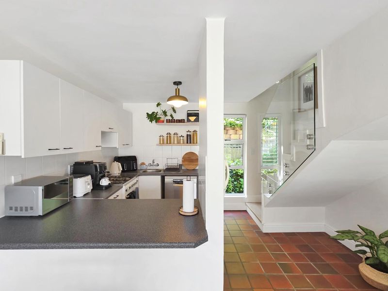 A functional kitchen featuring white cabinetry and dark countertops, adjacent to a tiled hallway with a modern glass balustrade staircase.