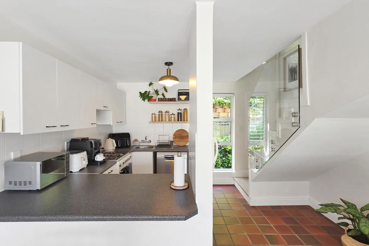 A functional kitchen featuring white cabinetry and dark countertops, adjacent to a tiled hallway with a modern glass balustrade staircase.