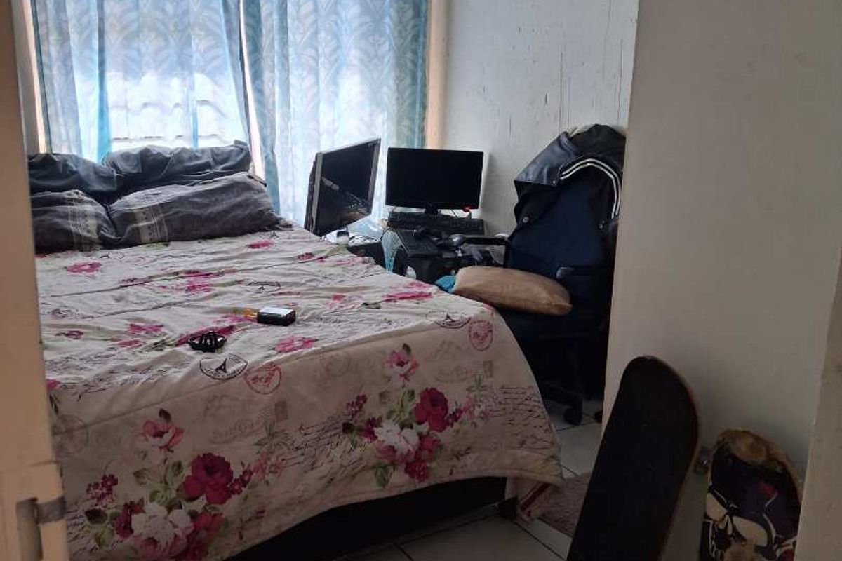 A compact bedroom featuring tiled flooring and a window with curtains, currently utilized as a sleeping and workspace area.