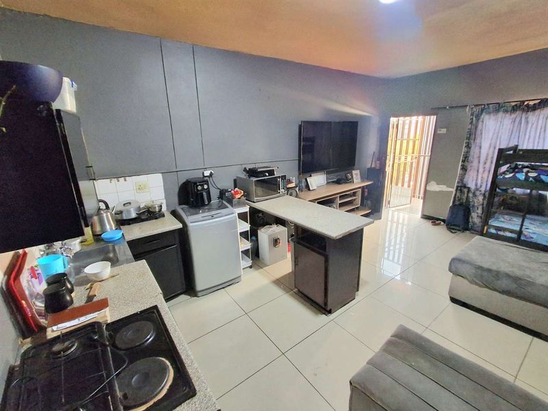 Open-plan living area featuring a compact kitchen with built-in cabinetry, gas hob, and tiled flooring, leading to an entrance with a security gate.