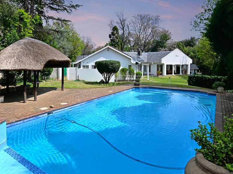 A well-maintained swimming pool area featuring a traditional thatched lapa, brick paving, and a view of the main house with a covered patio.