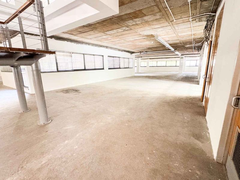 Spacious open-plan office area featuring an industrial aesthetic with exposed concrete ceilings, large windows, and a metal staircase.