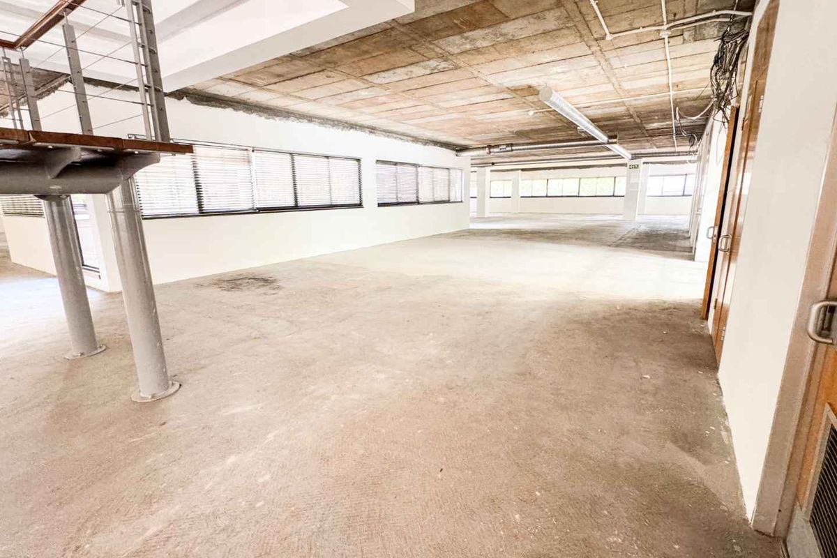 Spacious open-plan office area featuring an industrial aesthetic with exposed concrete ceilings, large windows, and a metal staircase.