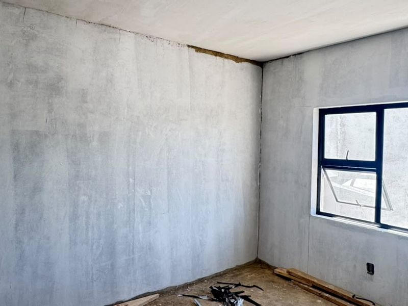 An unfinished bedroom interior featuring raw plastered walls, an installed aluminium window frame, and exposed floor surfaces, ready for final finishes and flooring.