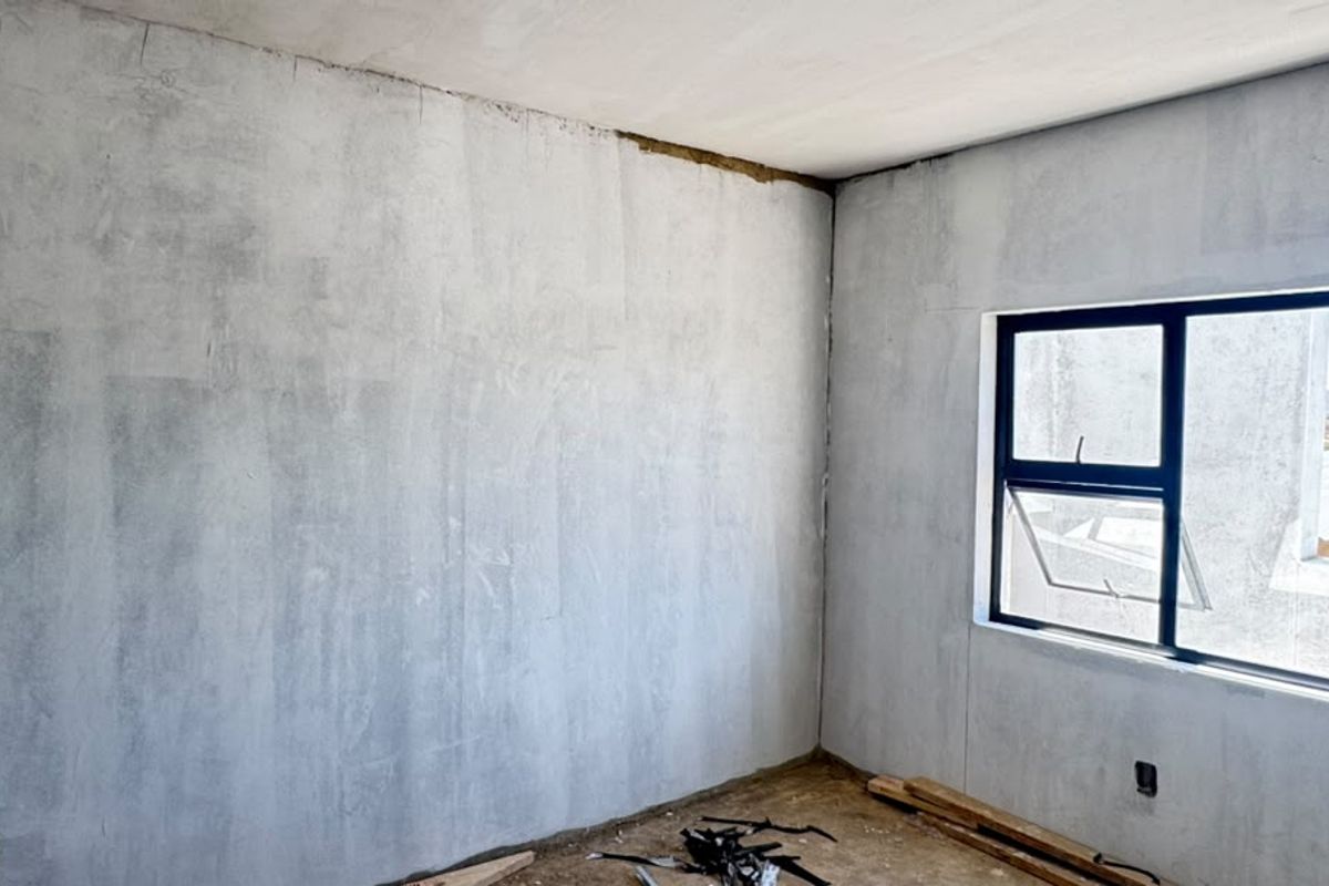 An unfinished bedroom interior featuring raw plastered walls, an installed aluminium window frame, and exposed floor surfaces, ready for final finishes and flooring.