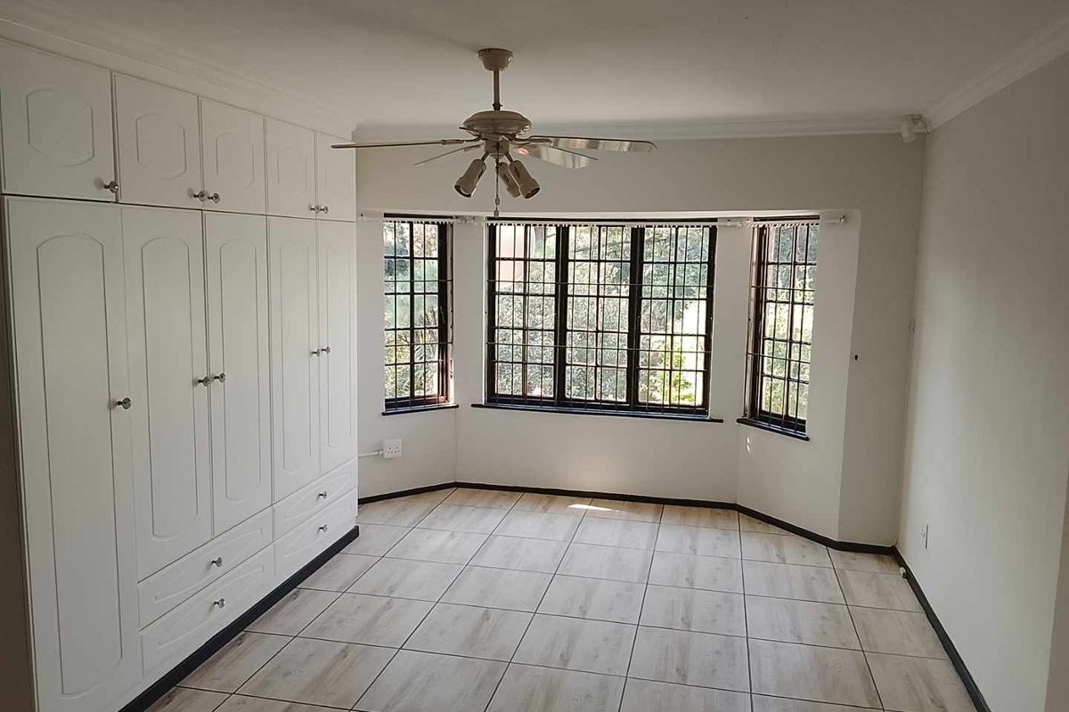 Spacious bedroom featuring large built-in cupboards, a ceiling fan, and a bright bay window with security bars and tiled flooring.