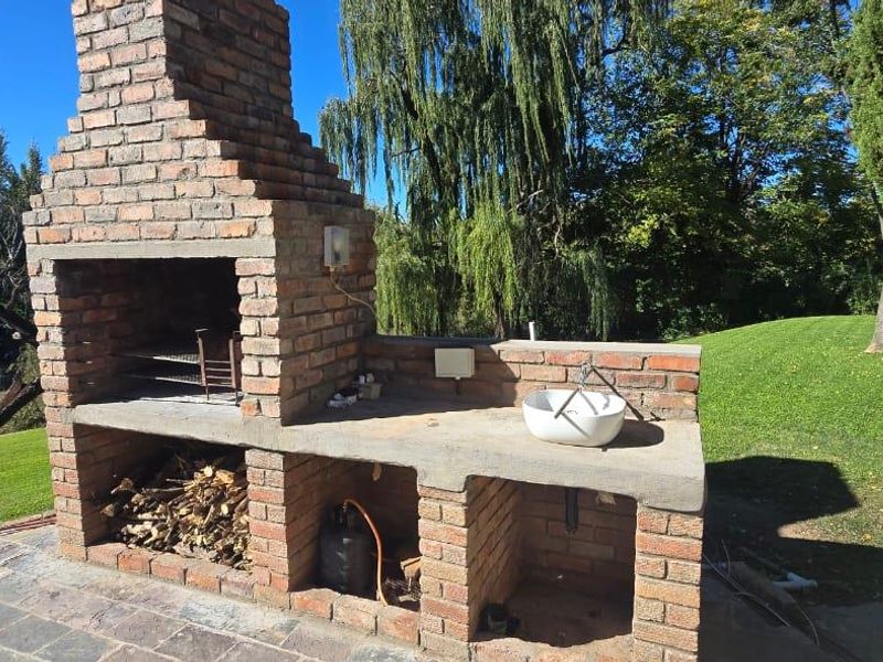 Outdoor patio area featuring a rustic brick built-in braai, concrete prep surface, and integrated sink, overlooking a lush garden and river view.