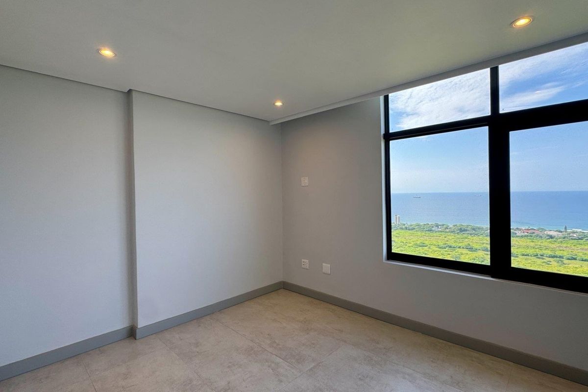 A bright, empty bedroom featuring modern tiled flooring, recessed downlights, and a large window offering expansive ocean and coastal views.