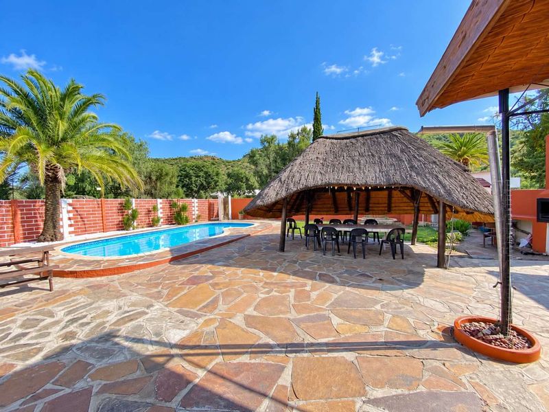 Outdoor entertainment area featuring a swimming pool, a traditional thatched lapa, and extensive stone paving, set against a brick boundary wall.