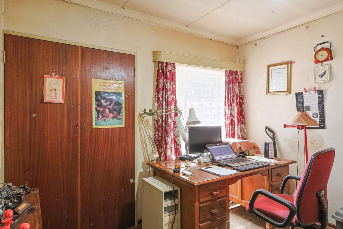 A bedroom featuring a built-in wooden wardrobe and a window, currently used as a home office space with dated wall finishes.