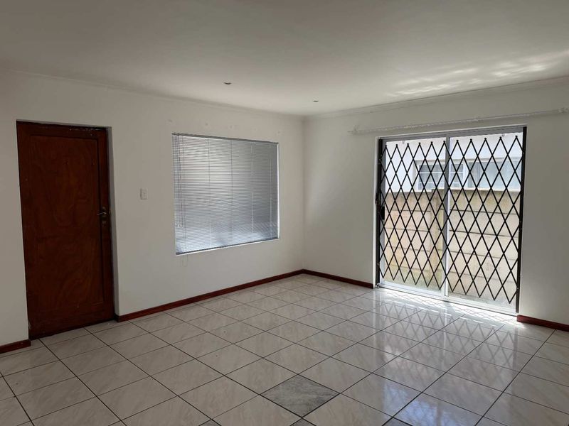 A bright, empty living area featuring neutral tiled flooring, a sliding glass door with security gate, and venetian blinds.