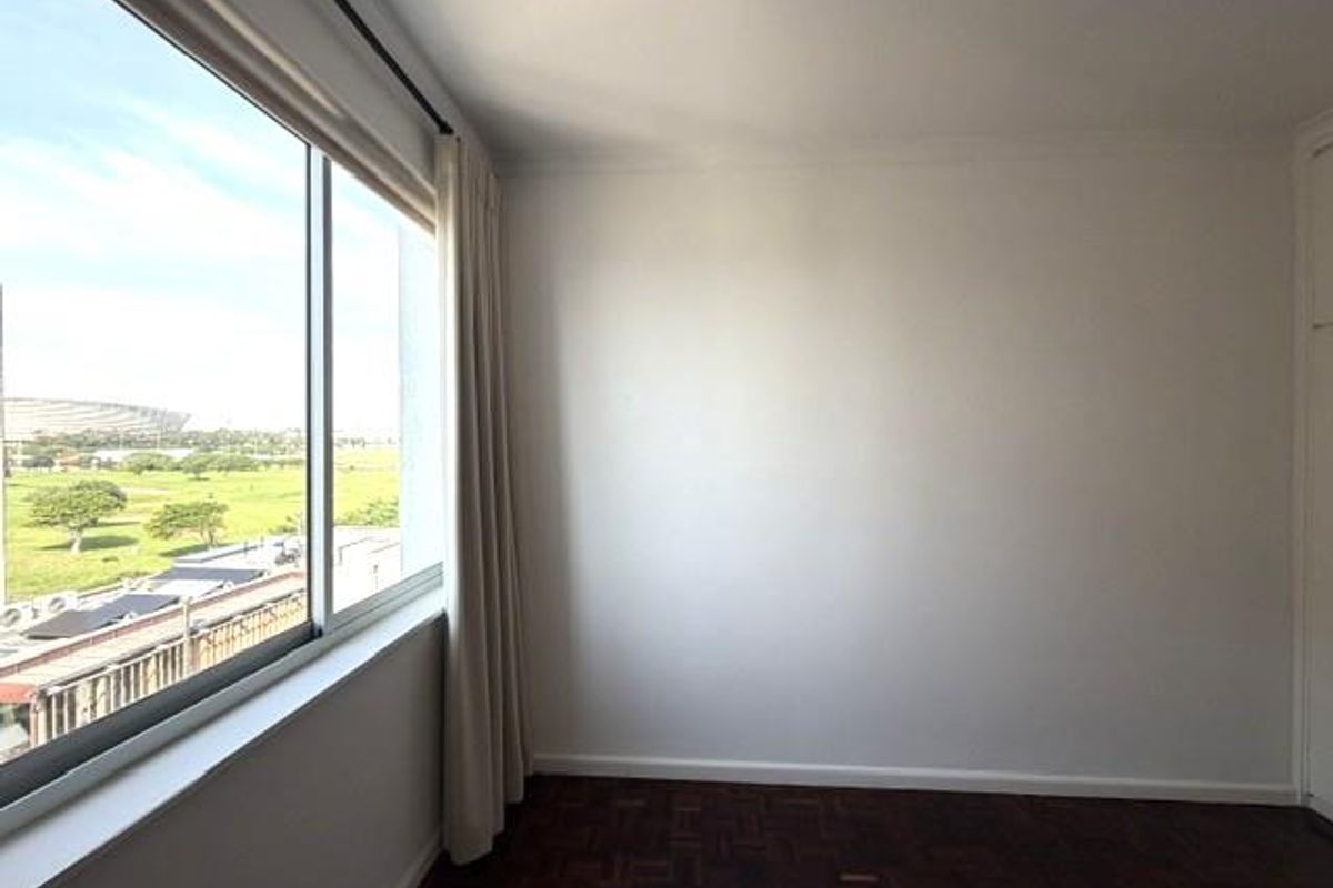 A bright, empty bedroom featuring classic parquet flooring, a large window with natural light, and built-in storage space.