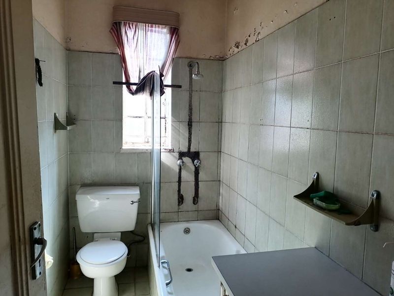 A dated bathroom featuring a bathtub with shower attachment, toilet, and tiled walls, currently requiring significant renovation and repair to the wall finishes.