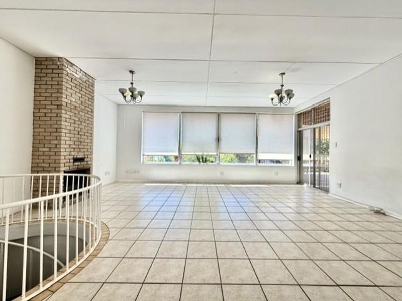 Spacious open-plan living area featuring a brick fireplace, tiled flooring, and large windows with roller blinds, leading to an outdoor patio area.